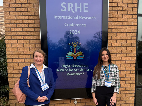 Foto: Prof. Liudvika Leišytė und Nina Bieling stehen vor einem Banner der SRHE Konferenz 2024