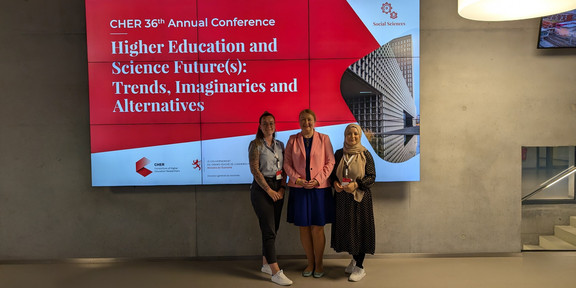 Gruppenfoto von Anja Westphal, Prof. Liudvika Leišytė und Sude Pekşen vor einem Banner der CHER Jahreskonferenz 2024