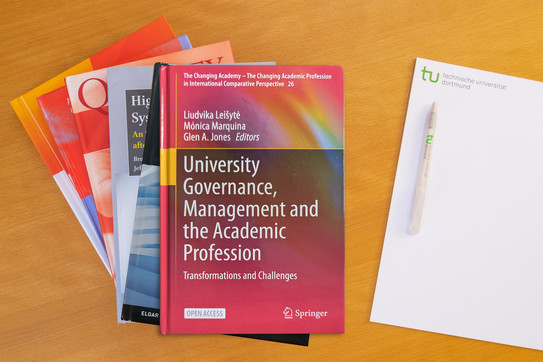 University governance, management and the academic profession Photo: Stack of books, to the right a notepad with a ballpoint pen on it