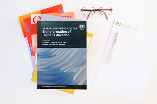 Photo: stack of books, the top book is the Research Handbook on the Transformation of Higher Education; next to it is a pad, a pen, and a pair of glasses