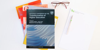 Foto: Bücherstapel, das oberste Buch ist das Research Handbook on the Transformation of Higher Education; daneben ein Block, ein Kugelschreiber und eine Brille