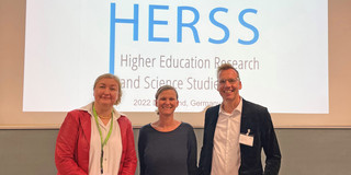 Gruppenfoto: Prof. Liudvika Leišytė, Prof. Tessa Flatten und Prof. Uwe Wilkesmann – im Hintergrund die Eröffnungspräsentation
