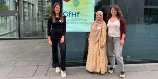 Group photo: Nina Bieling, Sude Pekşen and Anna-Lena Rose in front of a building, with a poster in the background displaying the logo of the German Society for Higher Education Research