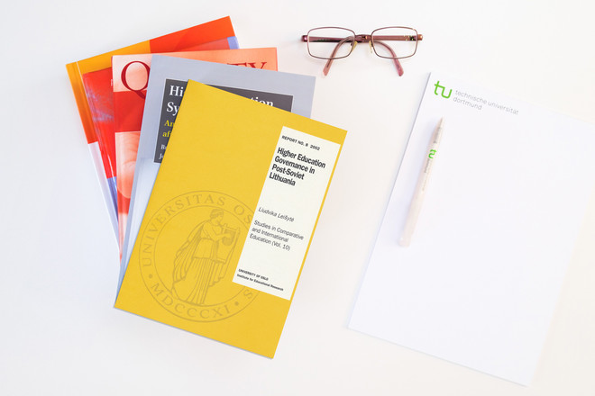 Bücherstapel, daneben ein Block, ein Kugelschreiber und eine Brille