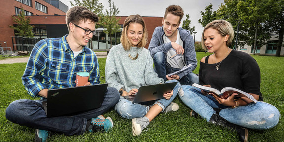 Four students are sitting on the grass and looking in a book.